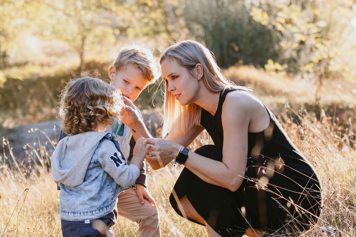 Blond Mother shares a beautiful moment wth her two young boys in the field, placing grass behind the one child's ear. 