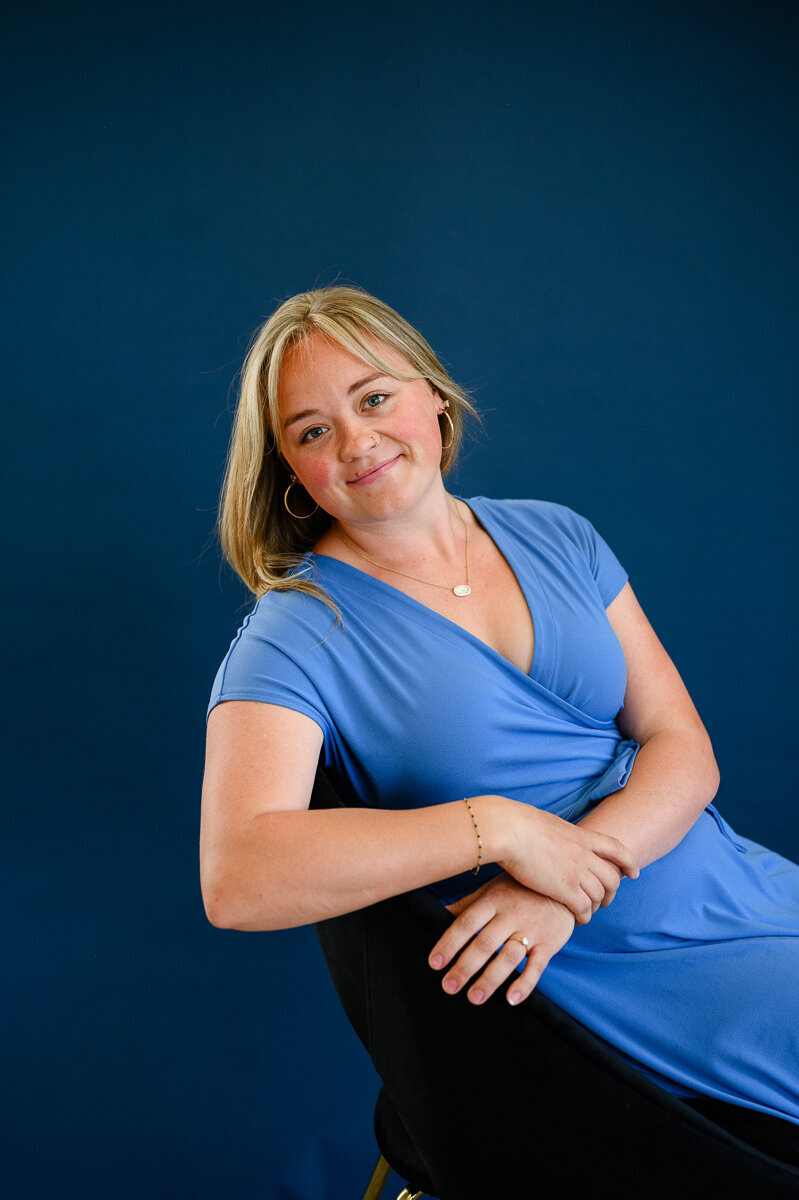 A brand photographer in Denver takes a photo of a woman business owner in a blue dress, sitting in a blue chair with a blue background for her brand photos during a branding photo shoot.