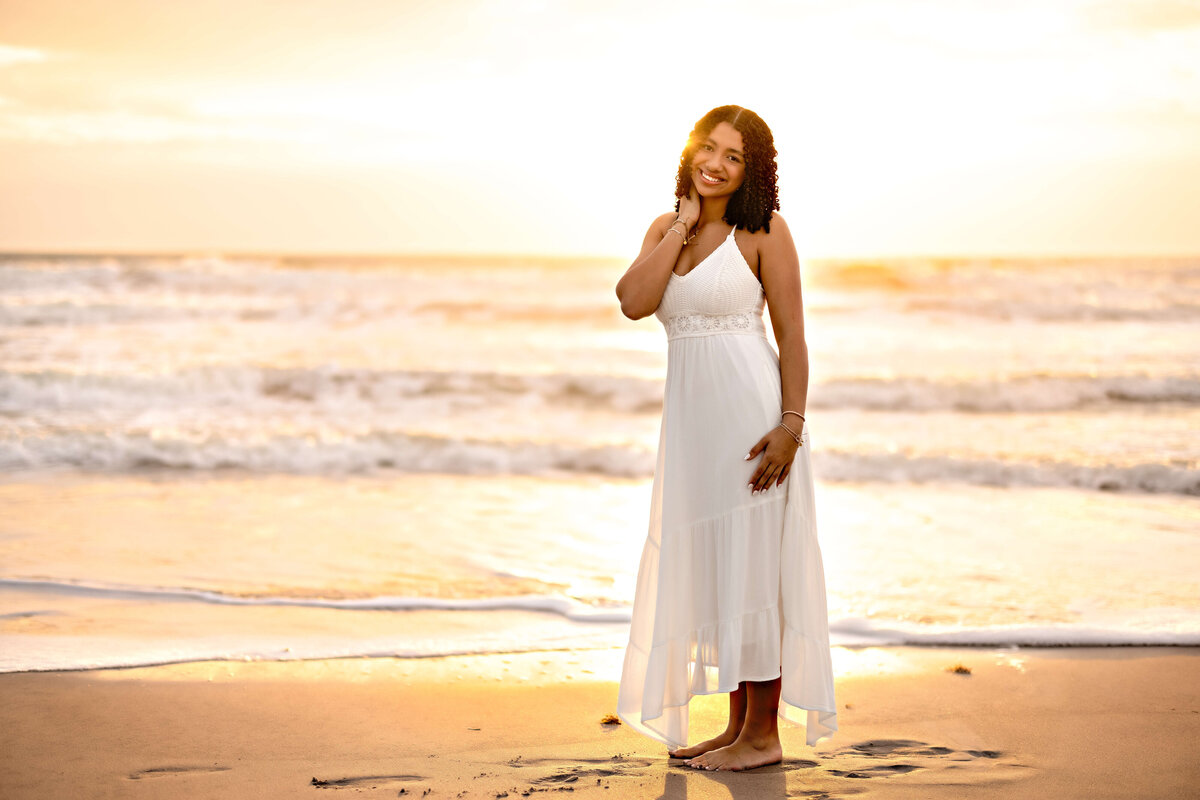 Orlando senior photographer capturing a golden hour portrait of a girl with one arm lifted behind her head, white dress glowing in beach light.