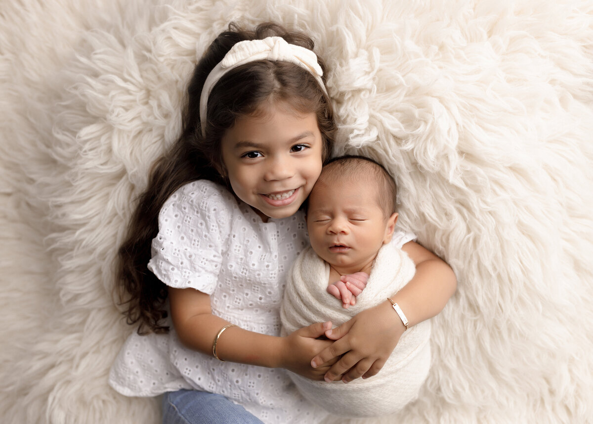 West Palm Beach and Boca Raton newborn photoshoot with baby brother and big sister. For the sibling newborn photo, the big sister and baby are atop of a fuzzy long faux fur rug. Big sister is smiling at the camera with the baby's head resting on her shoulder. Baby is sleeping in a white knit wrap with his hands peeking out the top. Aerial image.