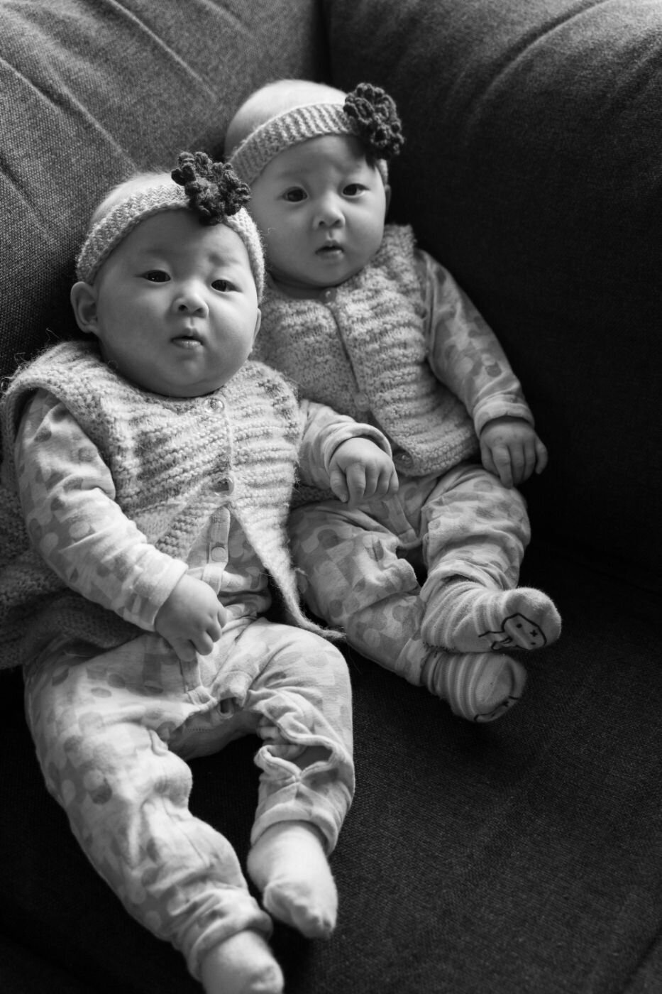 Black-and-white portrait of twin baby girls sitting side by side on a couch, wearing matching knitted vests and floral headbands.