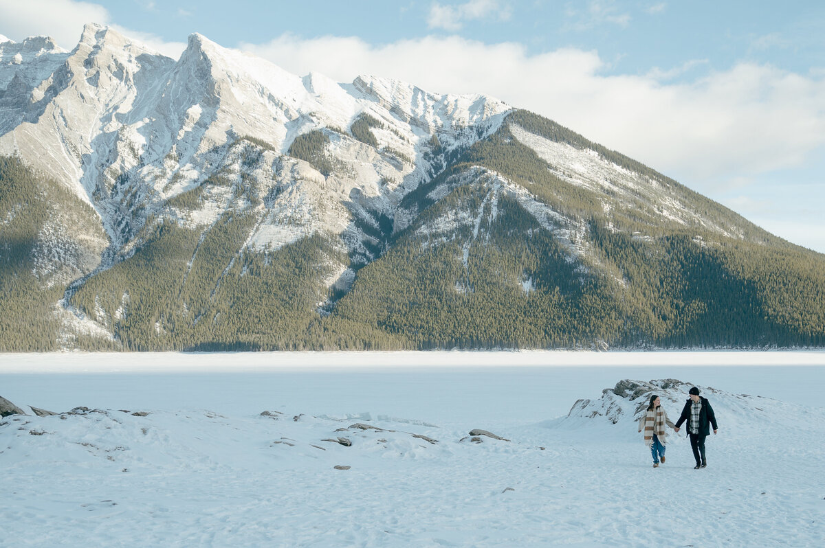 Couple walking across Lake Minnewanka during a winter engagement shoot in Banff - surrounded by mountain peaks.