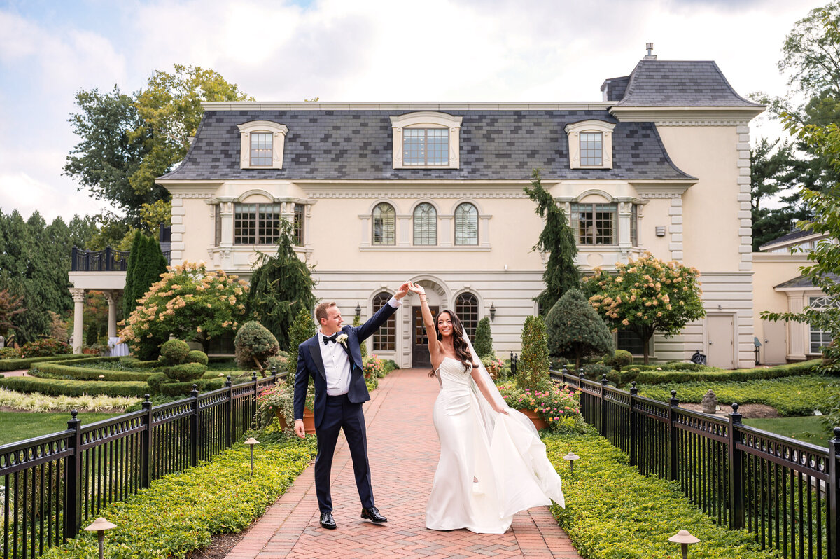 A bride and groom celebrate outdoors in front of a large, elegant building with manicured gardens and a brick walkway.