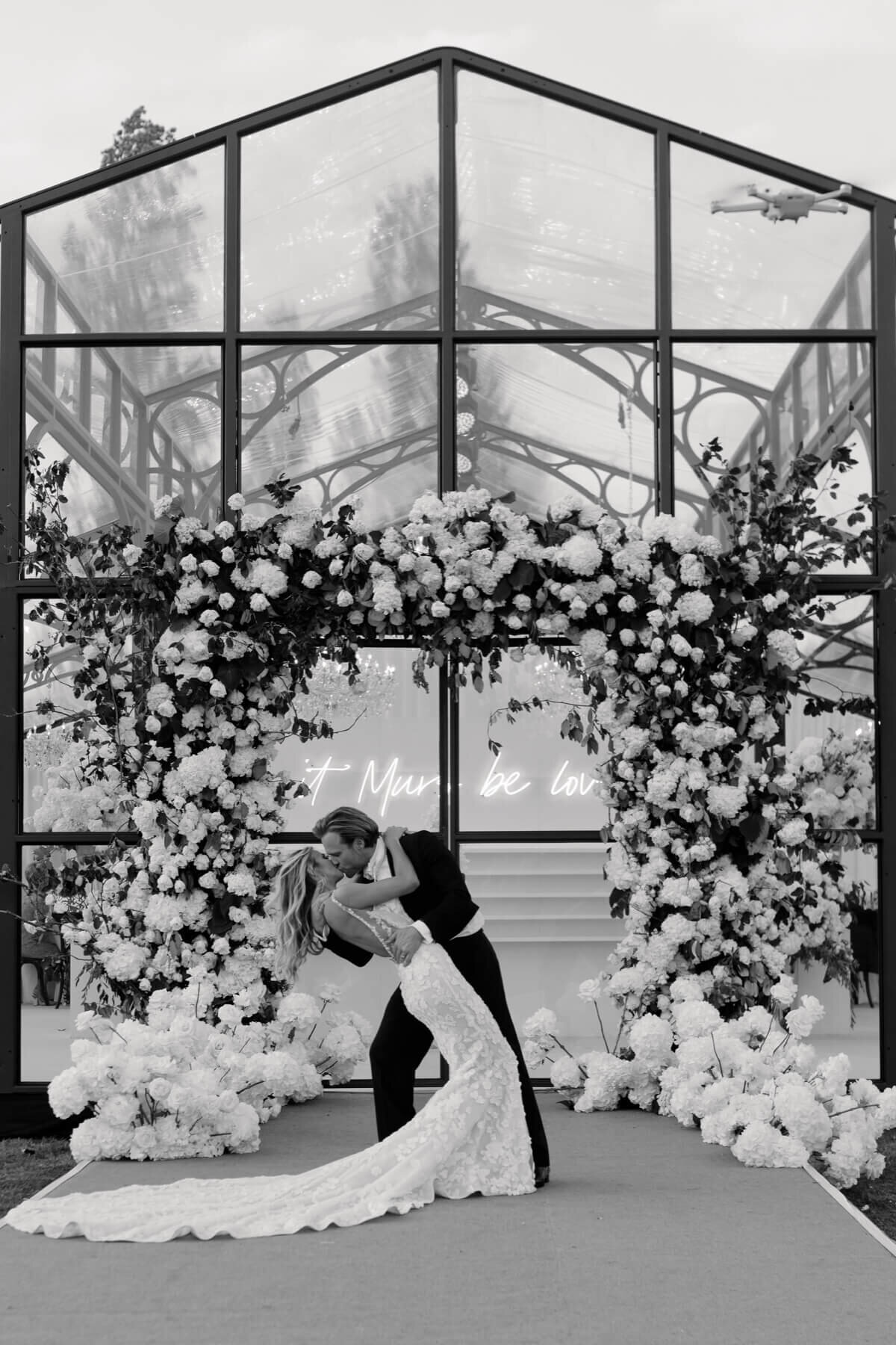 Amelia and Olly Murs in front of their glass wedding marquee with a stunning floral arch entrance.