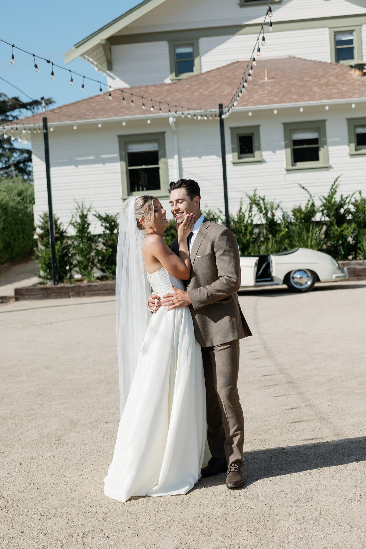 Couple embracing in front of a vintage car with their La Jolla wedding venue in the background.