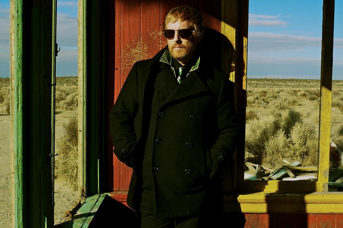 Male musician portrait The Mocking Bird standing in old shack in desert wearing sunglasses and dark overcoat