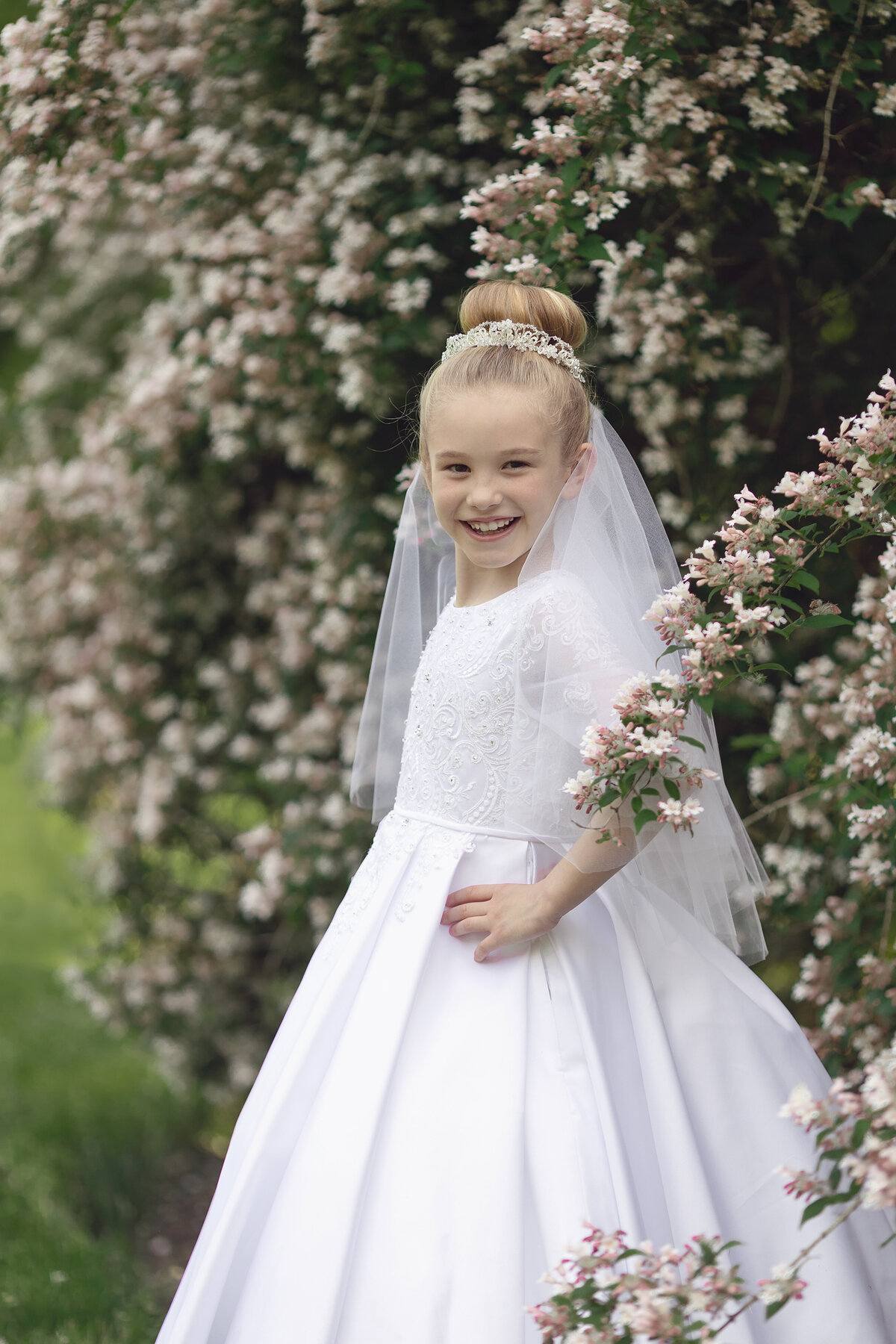 A young blonde girl smiles big with hands on her hips while standing for a New Jersey Communion Portrait Photographer among a white flowering bush