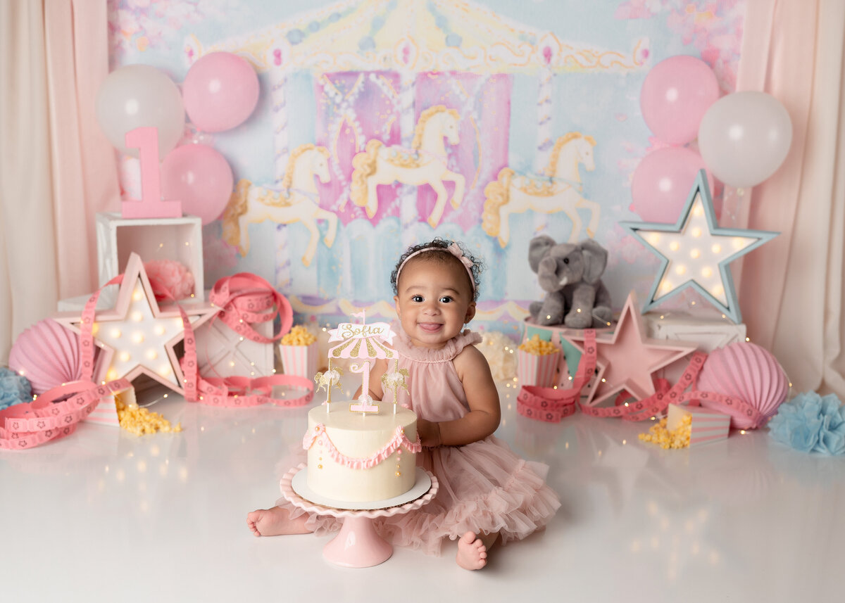 Carousel fairground cake smash. Baby in a pink tulle dress sits behind a carousel themed cake smiling at the camera. She hasn't touched the cake yet. In the background, there is a pale pink and pale blue carousel backdrop, popcorn, pink and white striped popcorn boxes, movie tickets, and pink and white balloons.