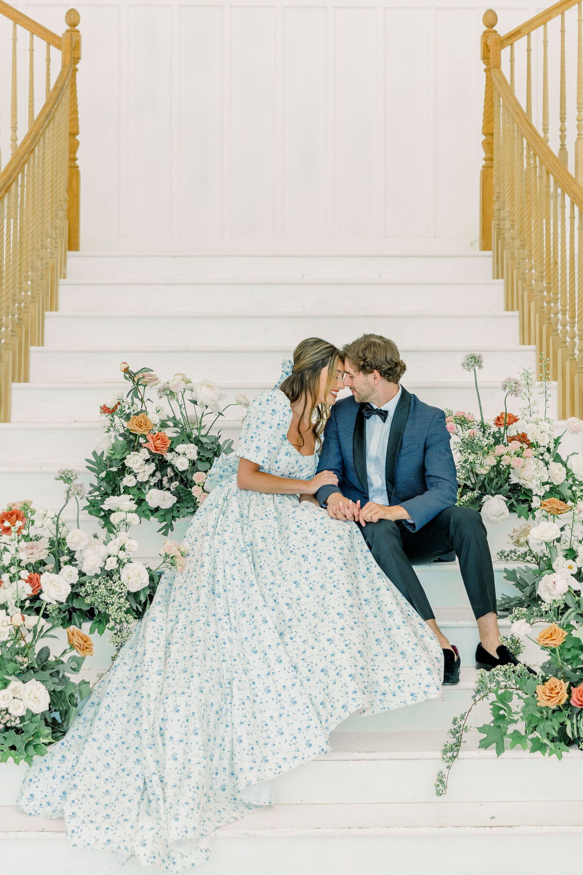 Ivy Rose Barn | A bride and groom sit on a white staircase at the Ivy Rose Barn in Rocky Mount, VA, surrounded by hundreds of florals. The bride wears a vintage-inspired floral gown with puff sleeves and a bow, while the groom is in a navy tuxedo.
