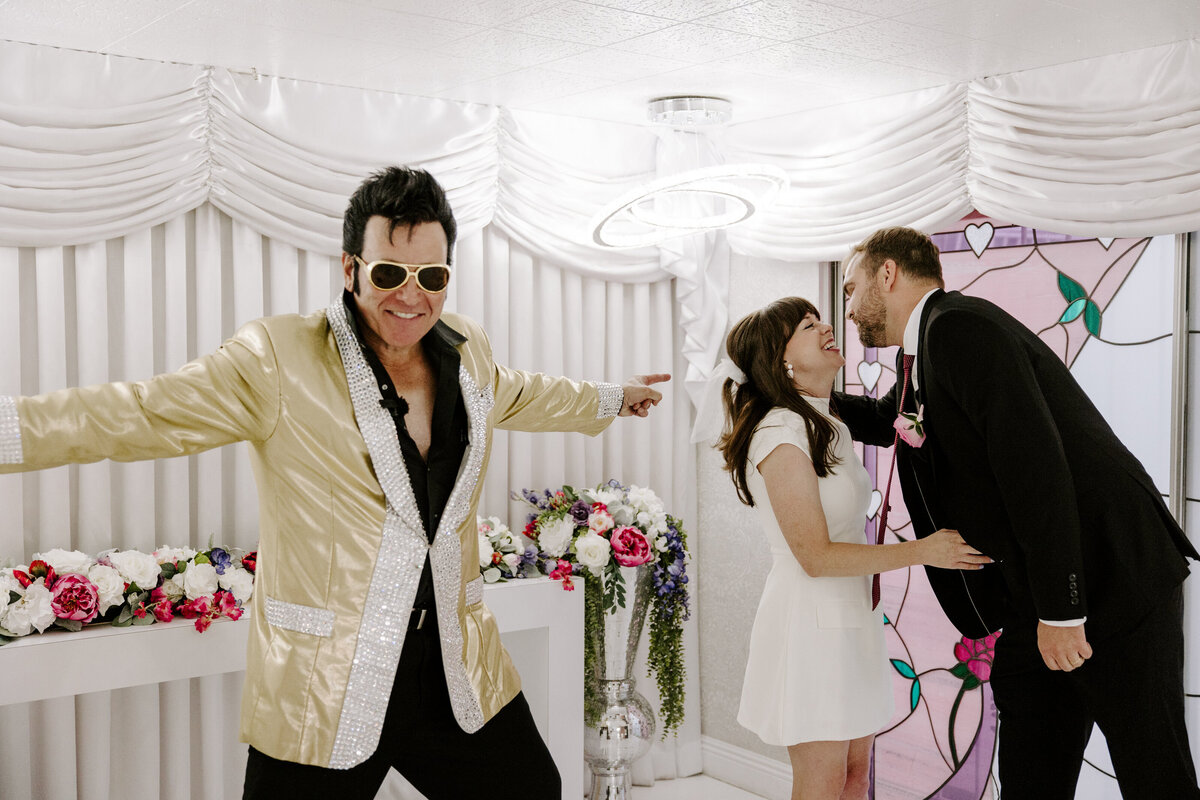 A couple leans in for a kiss during their elopement while an Elvis impersonator smiles and poses.