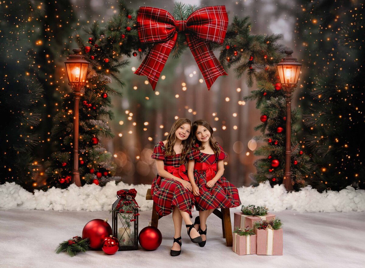 Two girls sitting on a bench with red plaid dresses, in front of a Christmas Arch and smiling at the camera.