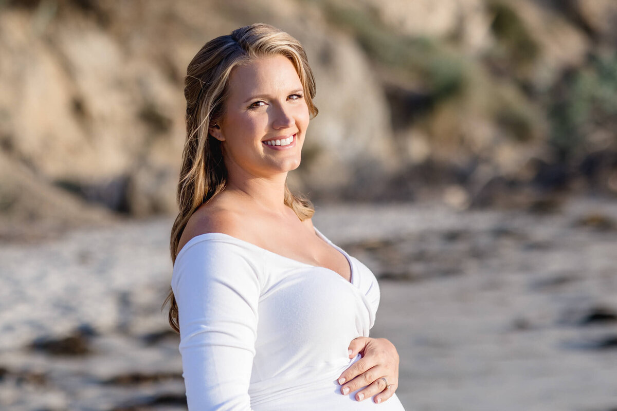 A woman with long hair, wearing a white off-shoulder top, smiles while standing on a sandy beach. She has her hand gently placed on her belly. The background features cliffs and soft-focus greenery.