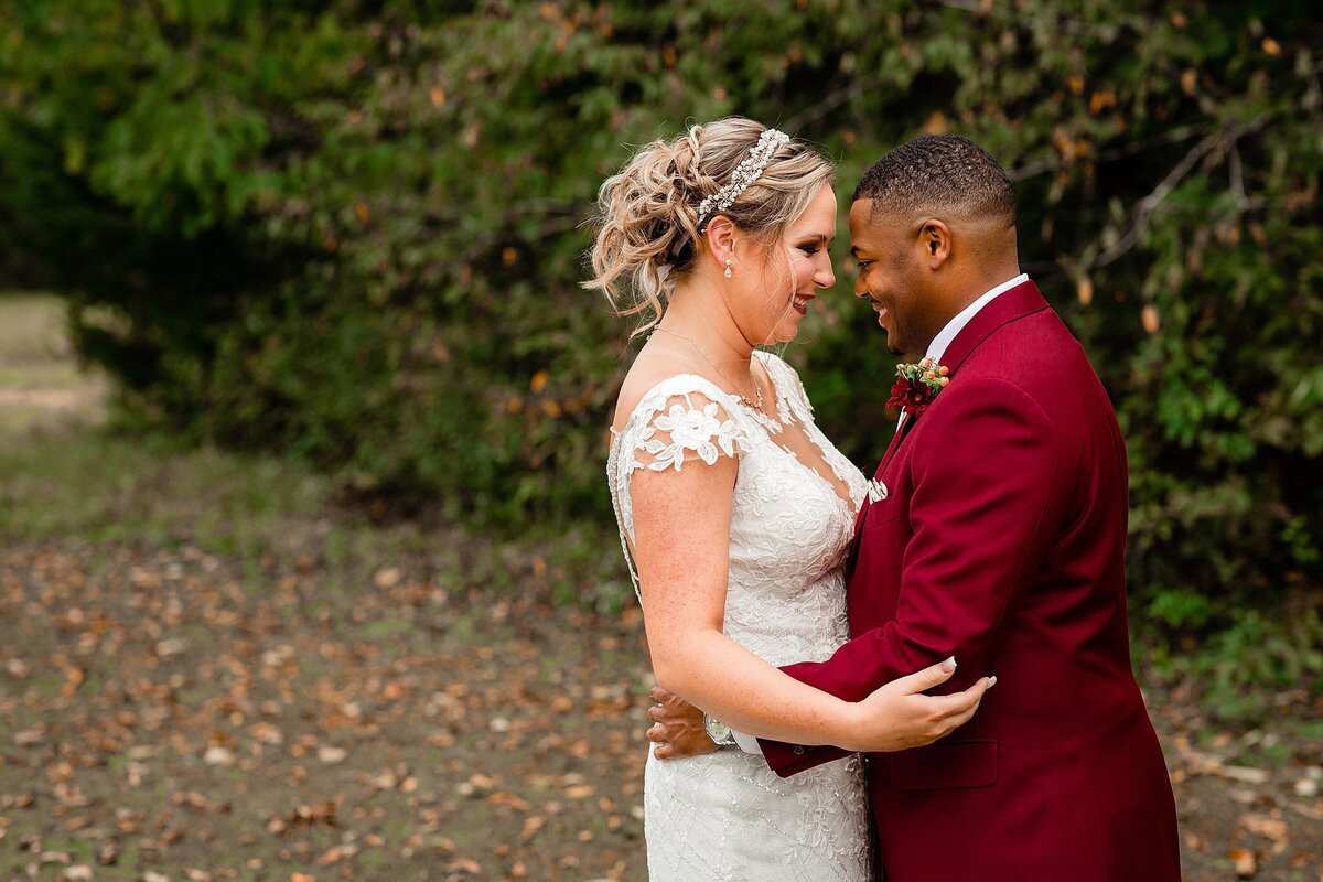 The bride wearing a lace wedding dress with a plunging neckline, lace sleeves and a rhinestone headband embraces the African American groom wearing a dark red suit with a rose boutonniere outside at Wild Turkey Ranch