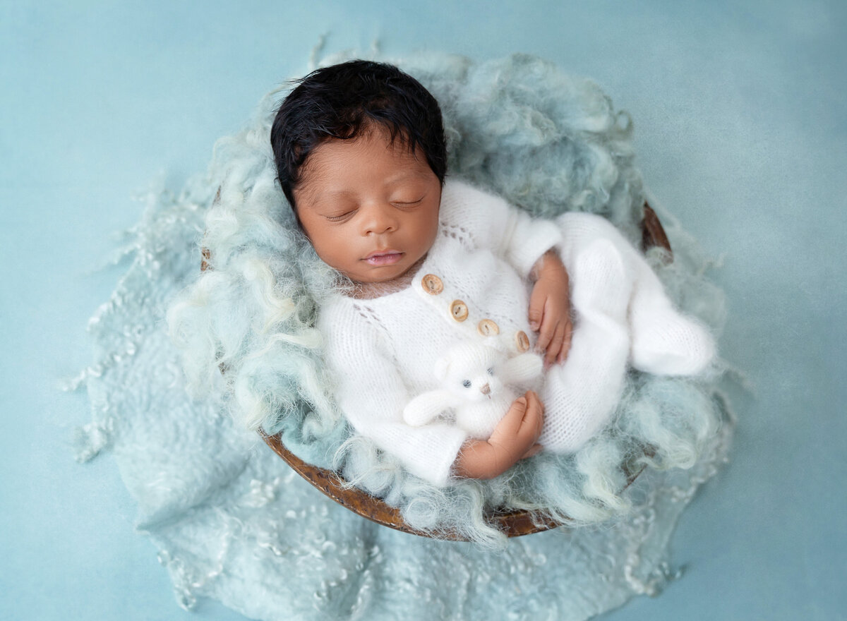 Baby boy in a white knit onesie sleeps atop of layered blue blankets for a newborn photoshoot.