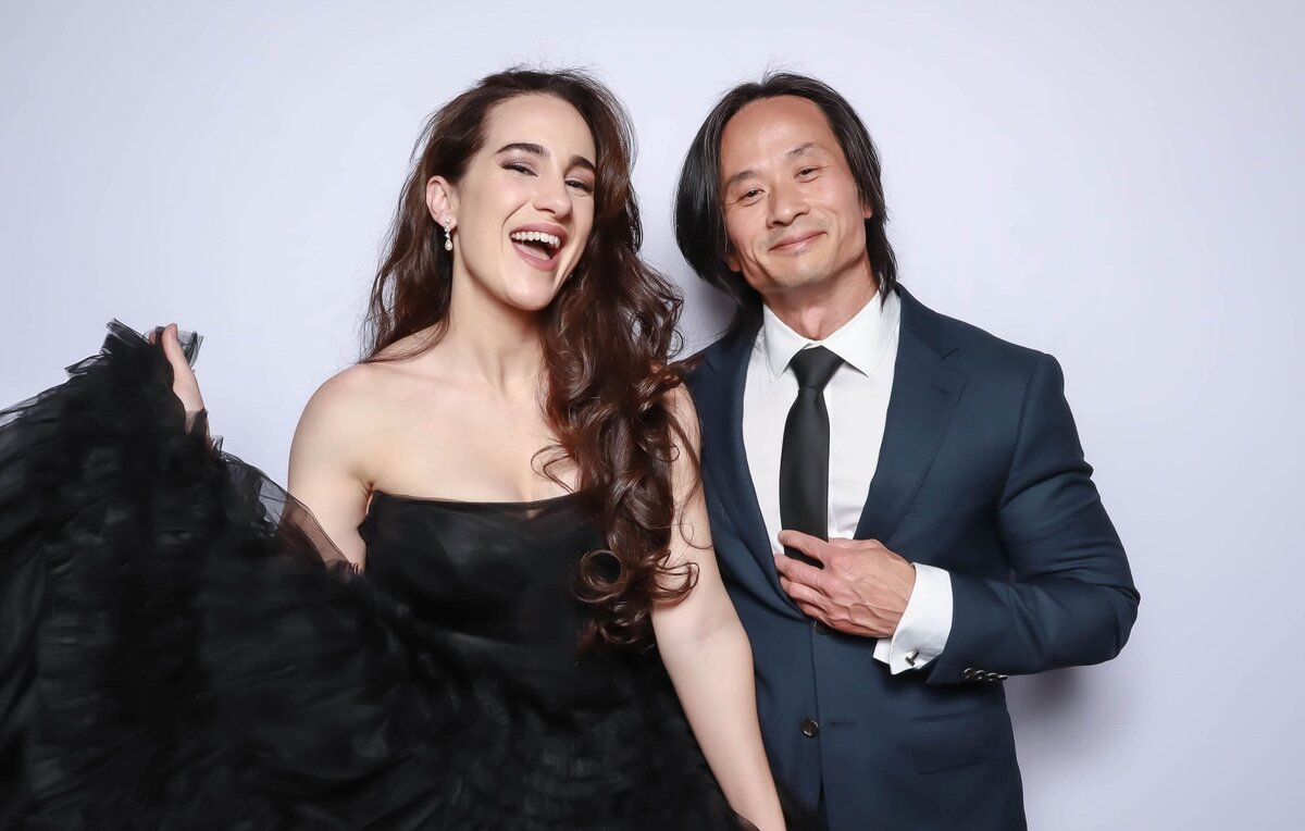 A woman in a strapless black gown and a man in a navy suit with a black tie pose together, smiling in front of a light backdrop at a formal event.