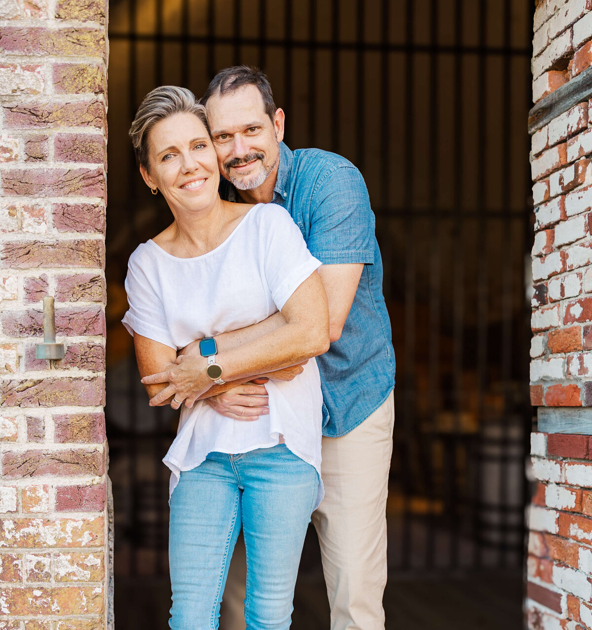 A couple stands in an open doorway, both smiling. The woman, in a white shirt and jeans, is in front, while the man, wearing a blue shirt and light pants, embraces her from behind. Framed by a brick wall in Fayetteville, NC, they radiate joy captured by their family photographer.