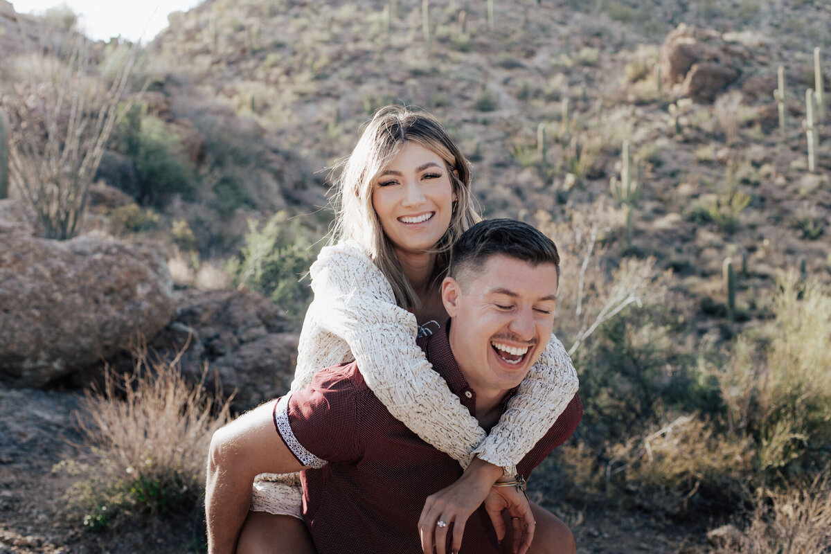 Couple having piggy back ride and laughing with desert landscape in background during golden hour in Phoenix, Arizona.