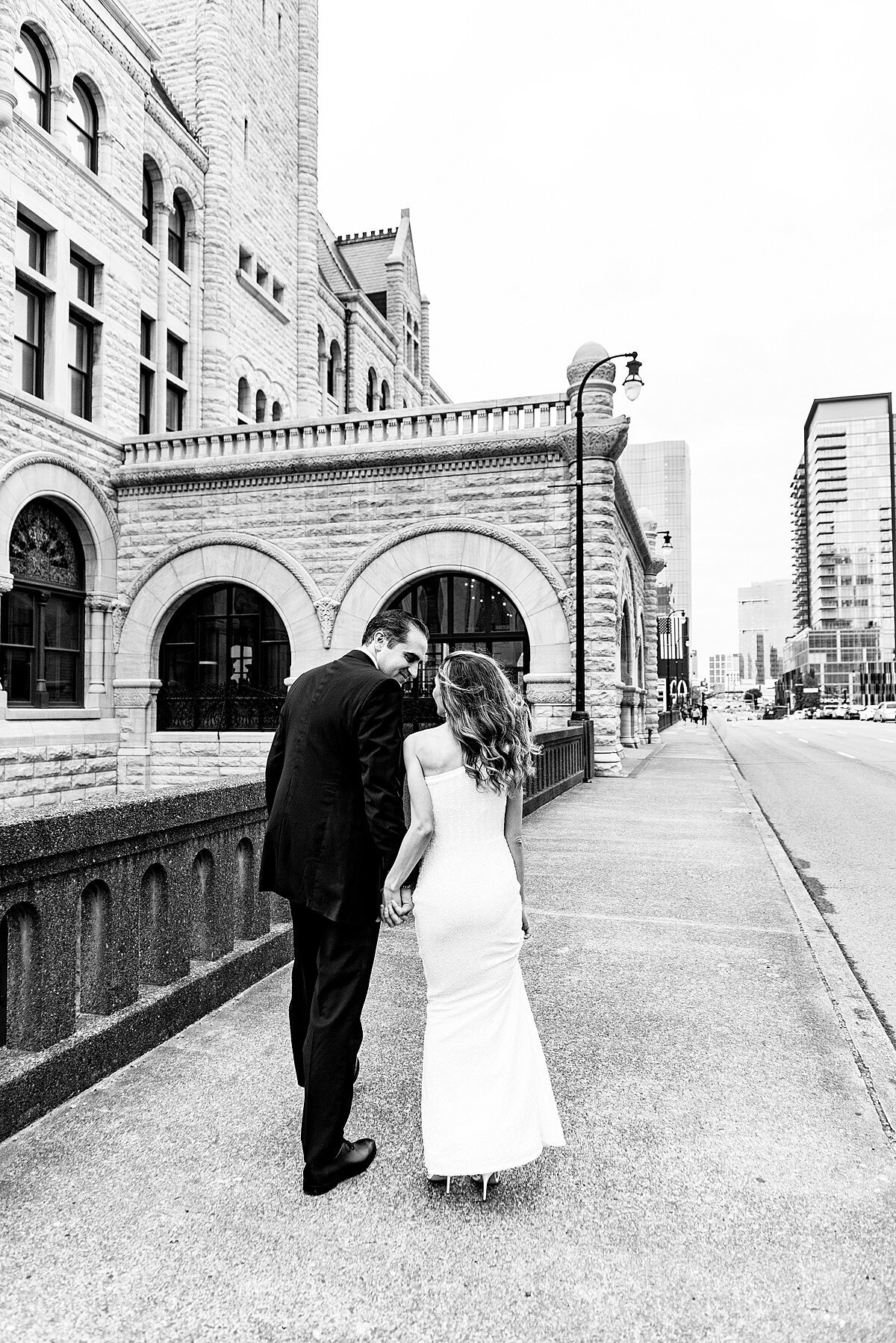 A black and white image of the bride and groom walking away while gazing up at each other holding hands in front of the Historic Union Station in Nashville. The bride is wearing a one shoulder sheath dress and the groom is wearing a black tuxedo.