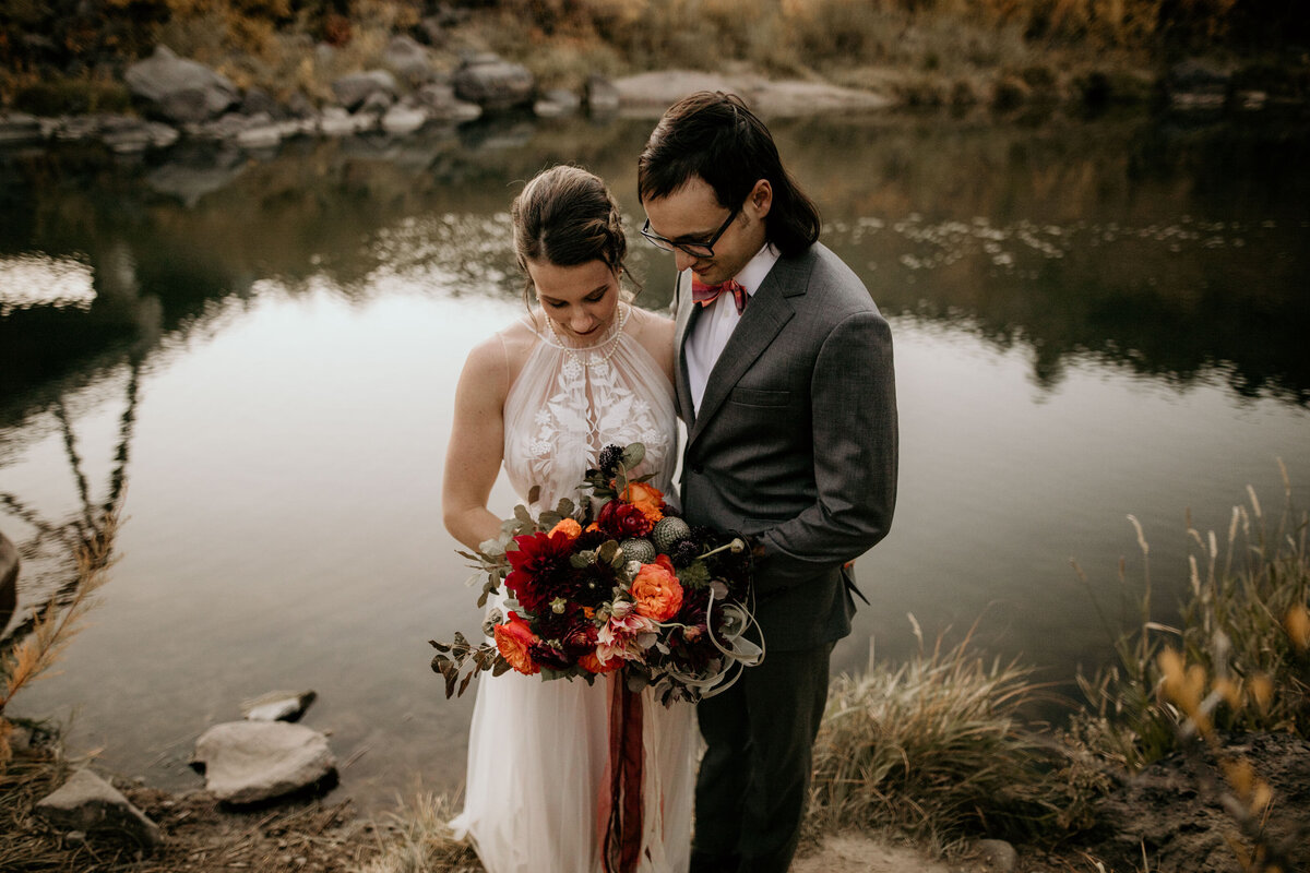 bride and groom looking at bouquet by a river