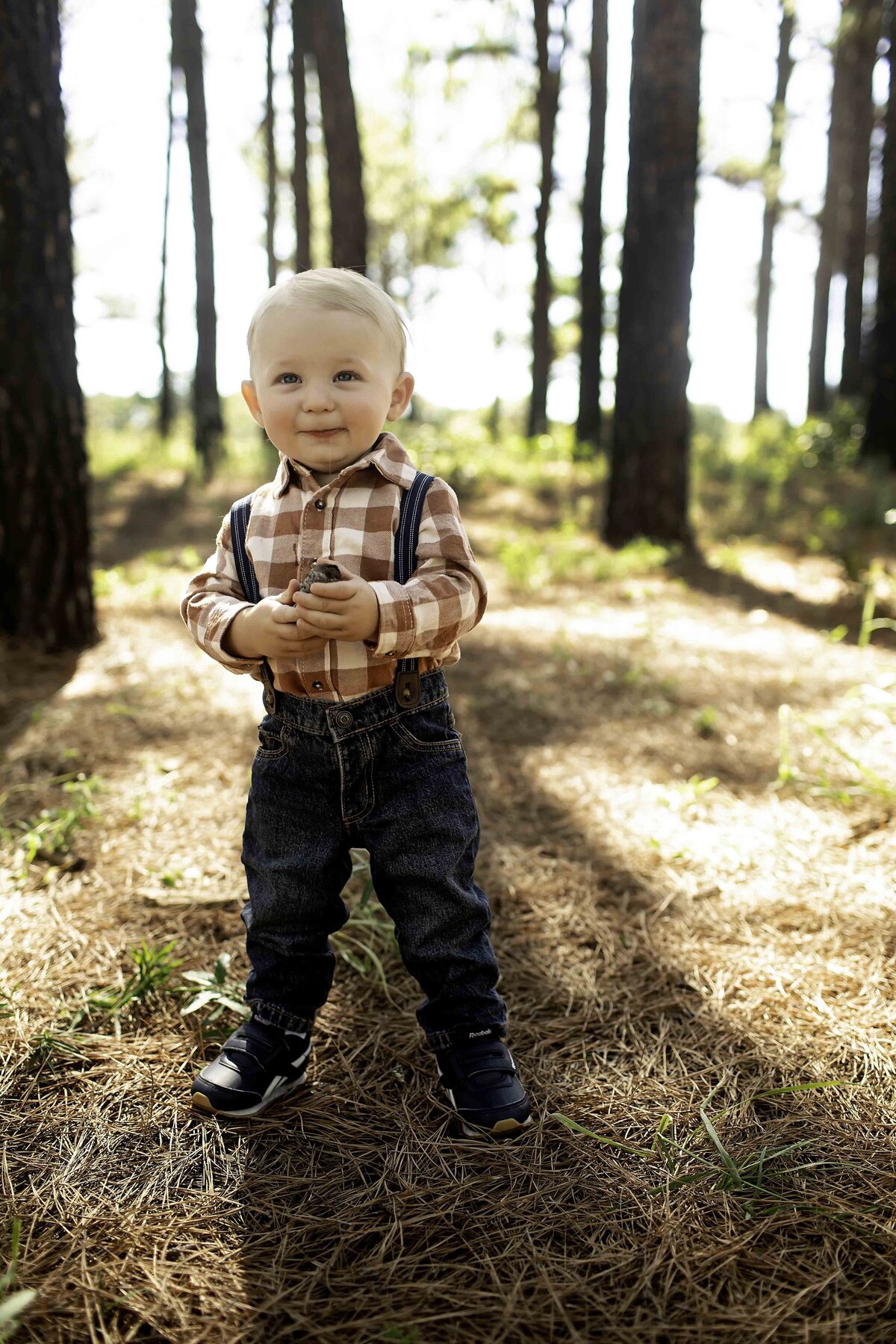 Birthday boy wearing suspenders and overalls, standing under the pine trees with a playful expression