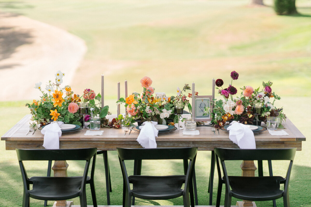 Orange, deep red, white, flower blooms with greenery on a farm table set up outside in a field