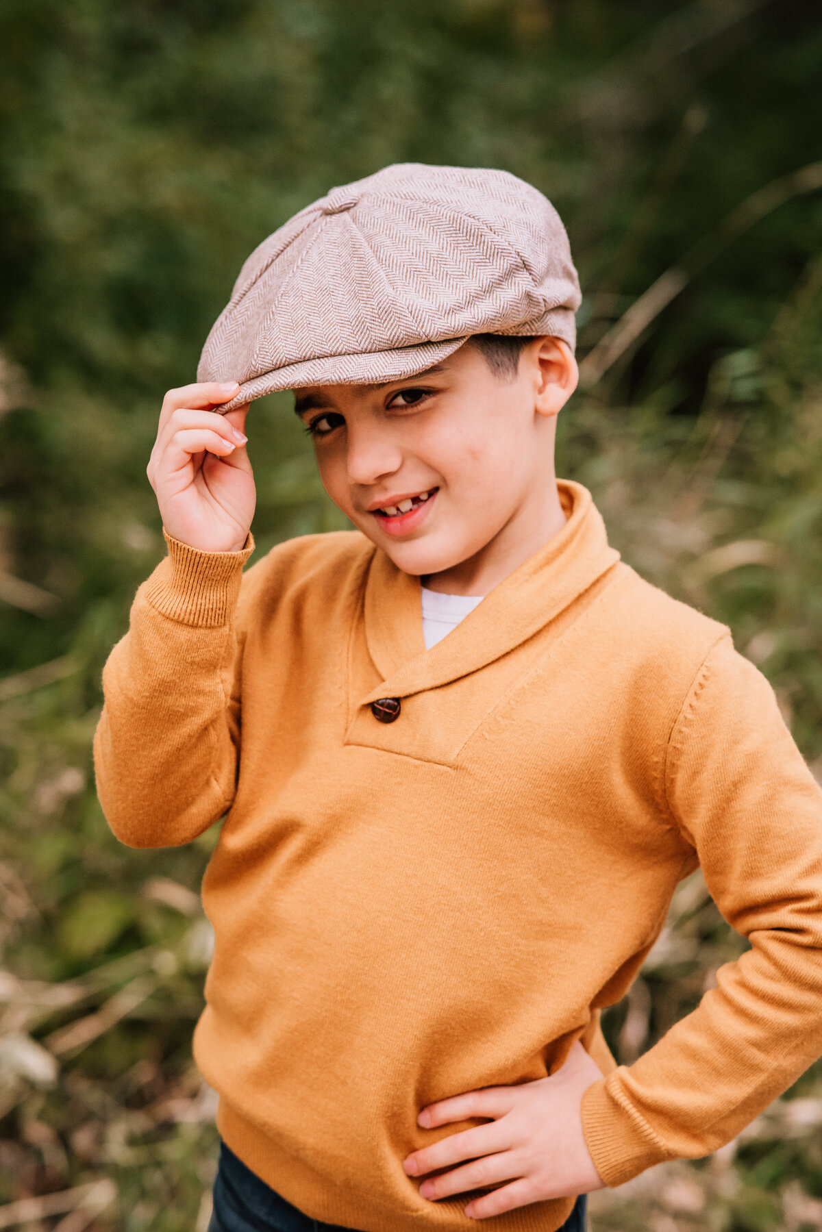 A young boy in a mustard yellow sweater and newsboy cap strikes a playful, confident pose, tipping his cap slightly as he smiles at the camera.