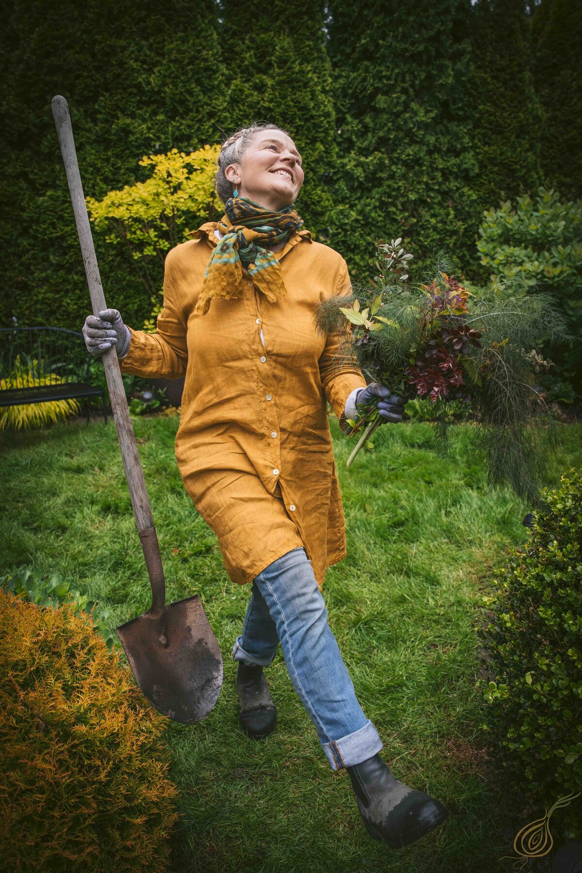 A woman smiling, wearing a yellow shirt and jeans stands holding a shovel, ready for gardening.