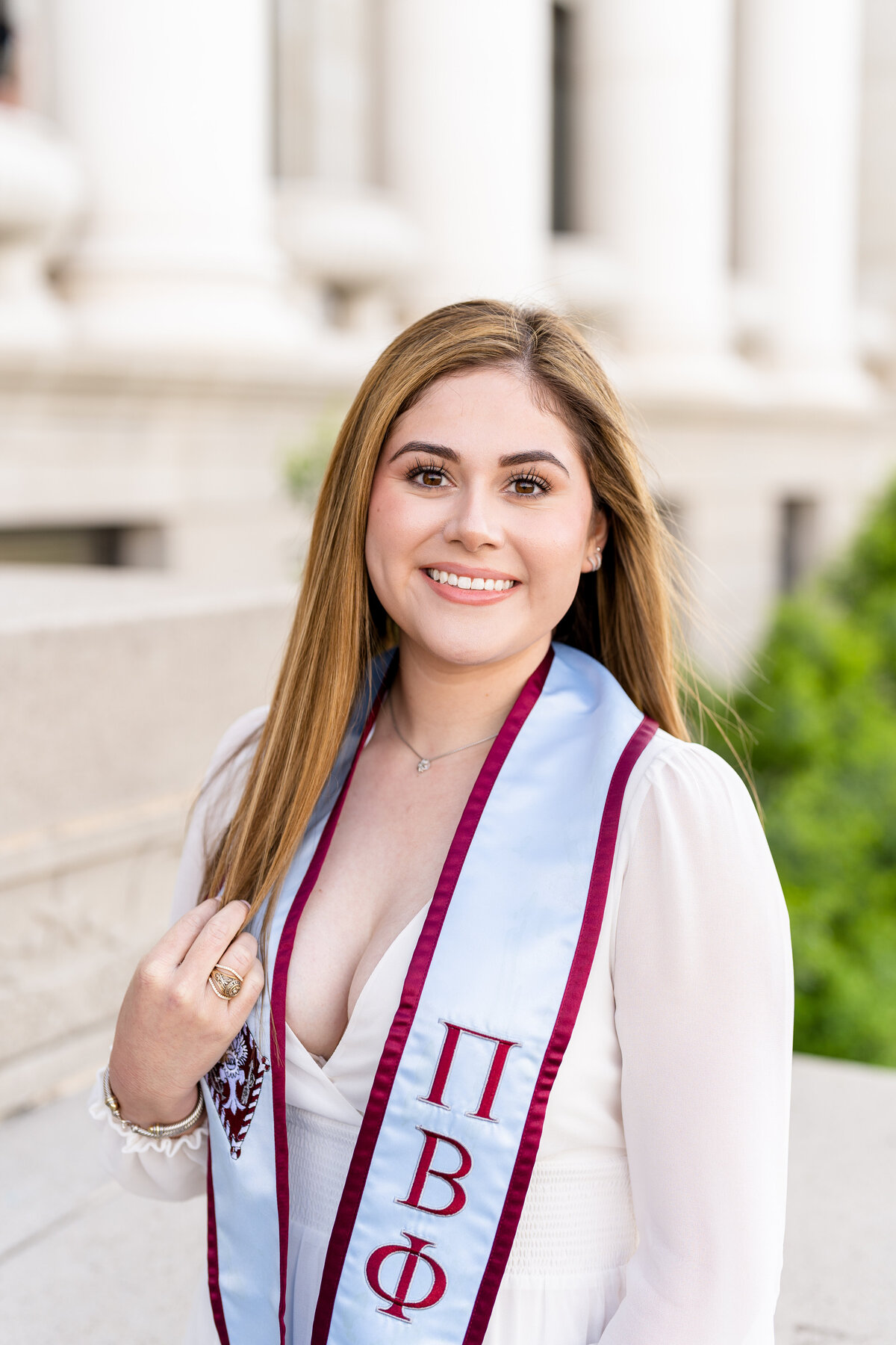 Texas A&M senior girl wearing Pi Phi stole and touching hair while smiling and sitting in front of Administration Building