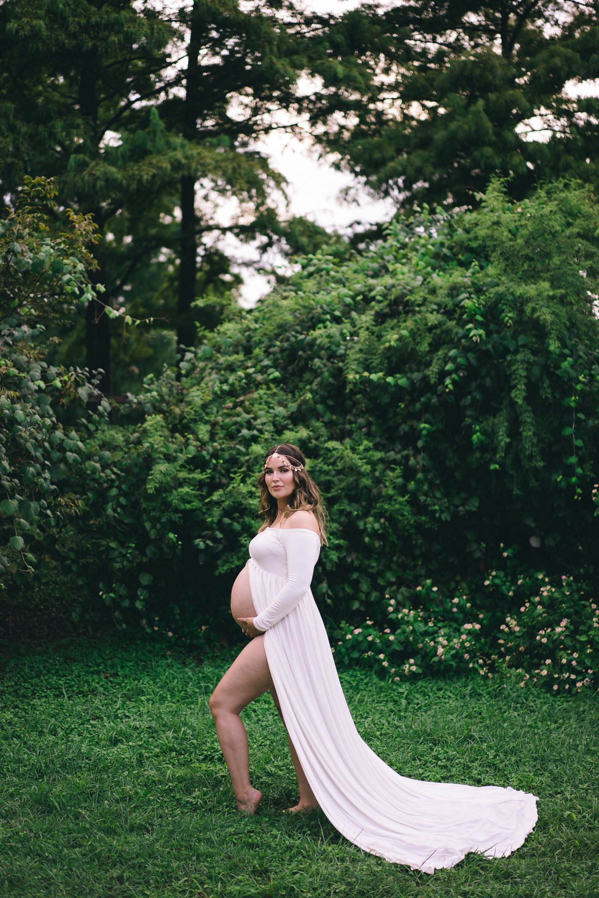 Woman in luscious green field posing for her maternity photography photo shoot in a white dress and is flowing in the wind.