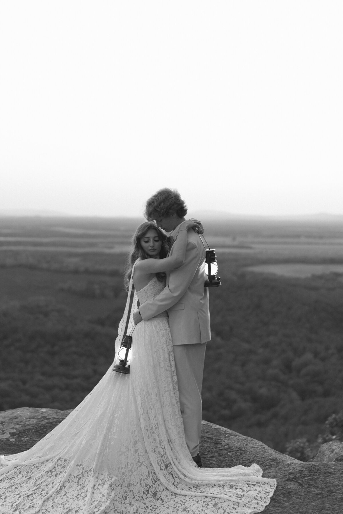 an elopement photo of a couple standing on a cliff at CC Overlook in Petit Jean State Park, Arkansas while holding lanterns during blue hour.