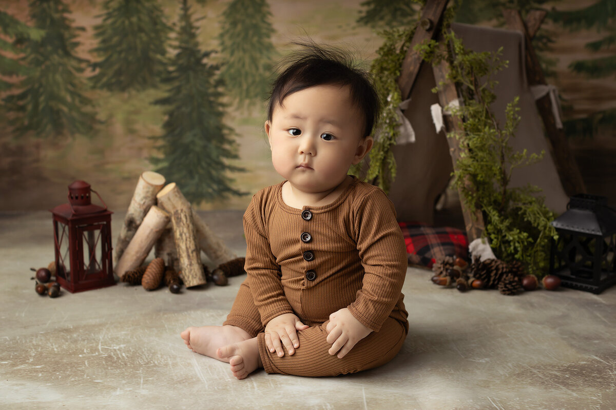 A sweet baby dressed in a cozy, buttoned romper sits on a rustic-themed set decorated with pine logs, lanterns, and a small tent against a forest backdrop. This charming milestone photo by a Dallas baby photographer beautifully captures a sense of exploration and curiosity, perfect for celebrating this precious stage of growth.