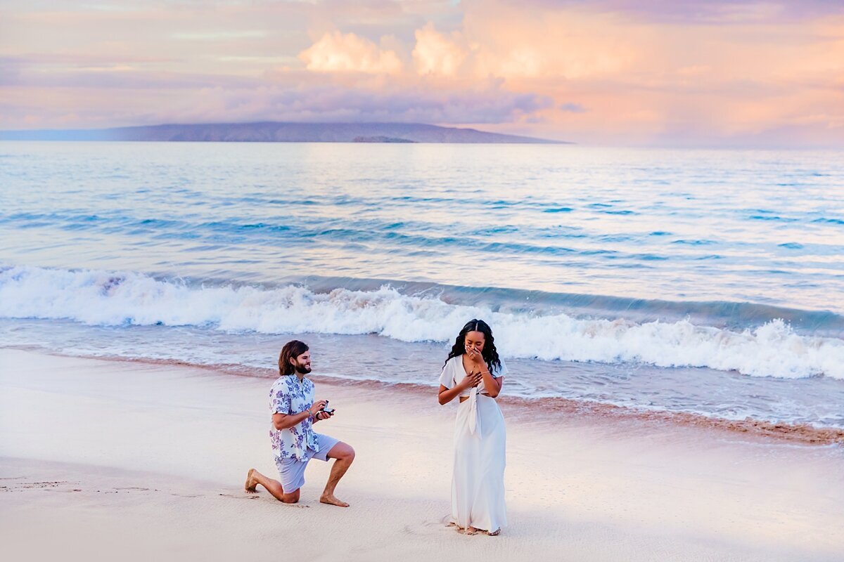 Woman in a white dress covers her mouth on the beach in happy surprise as her partner gets down on one knee to propose
