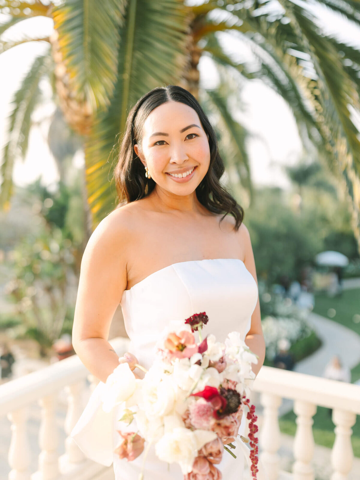A joyful bride in a strapless white dress holds a vibrant bouquet of pink and white flowers.