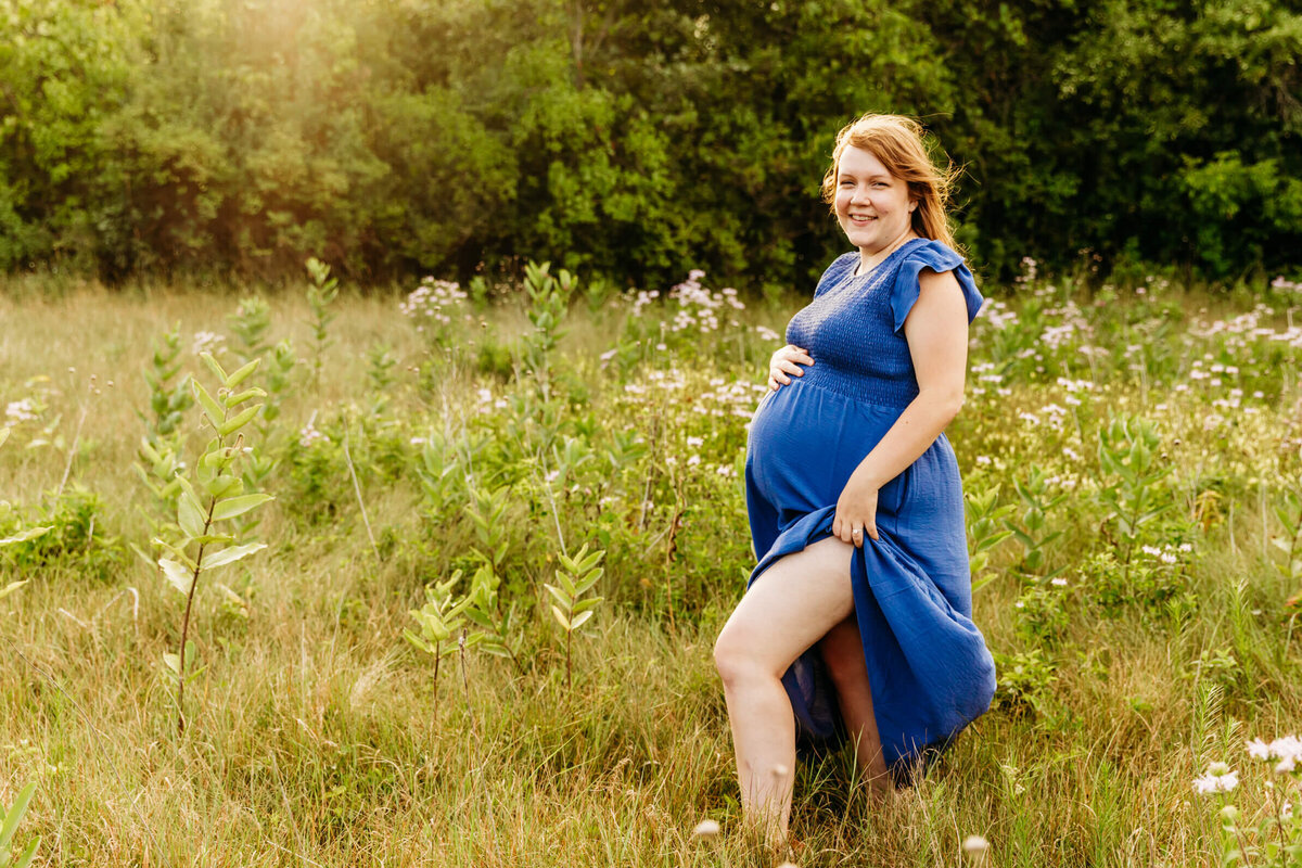 expecting mama in a blue dress standing in a wildflower field at sunset and smiling