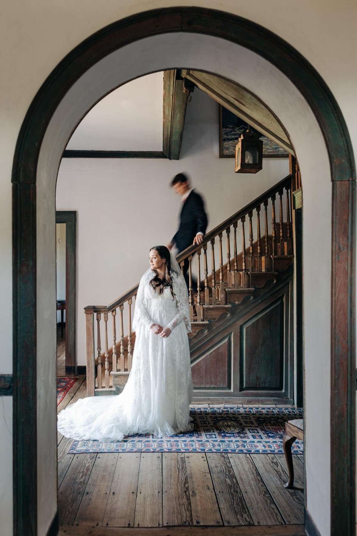 A bride standing in an archway with a groom descending a staircase behind her.