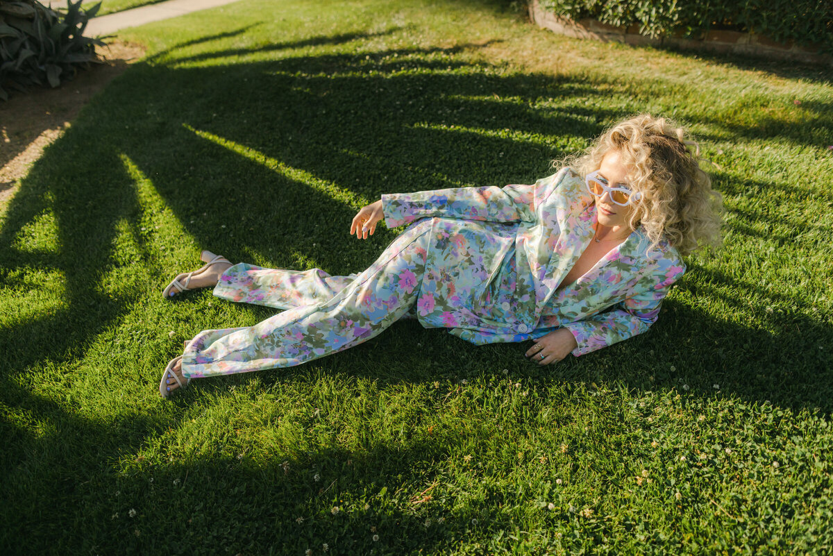 A fashion editorial moment sprawled in the grass — pastel floral suit, strappy heels, and retro sunglasses casting shadows with attitude. Greenville SC, Dani Lacey.