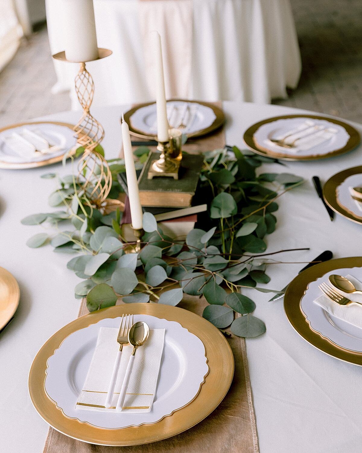white table with gold charger and white scalloped edge plate with a napkin and white and gold flatware in the center. The table has a white linen with a gold table runner. The centerpiece is a stack of antique books surrounded by eucalyptus and white candles in gold candlesticks.