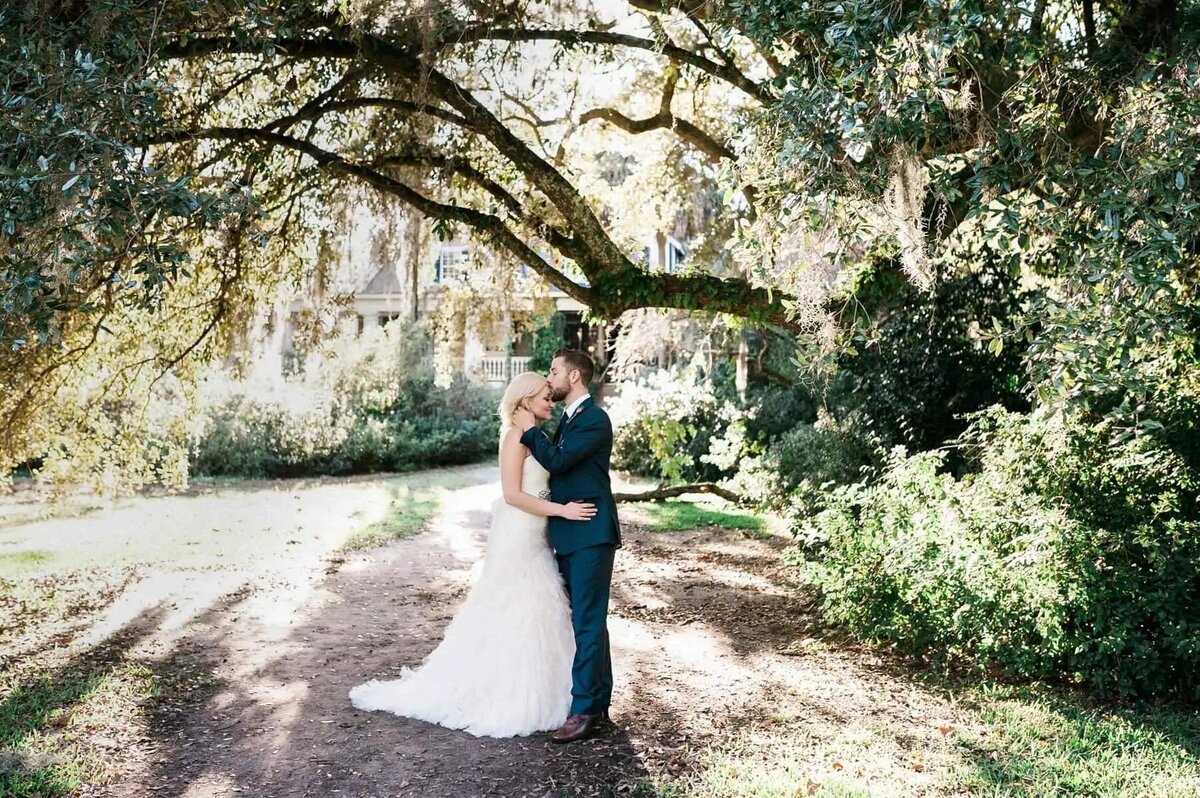 A groom kissing a brides head under a tree.