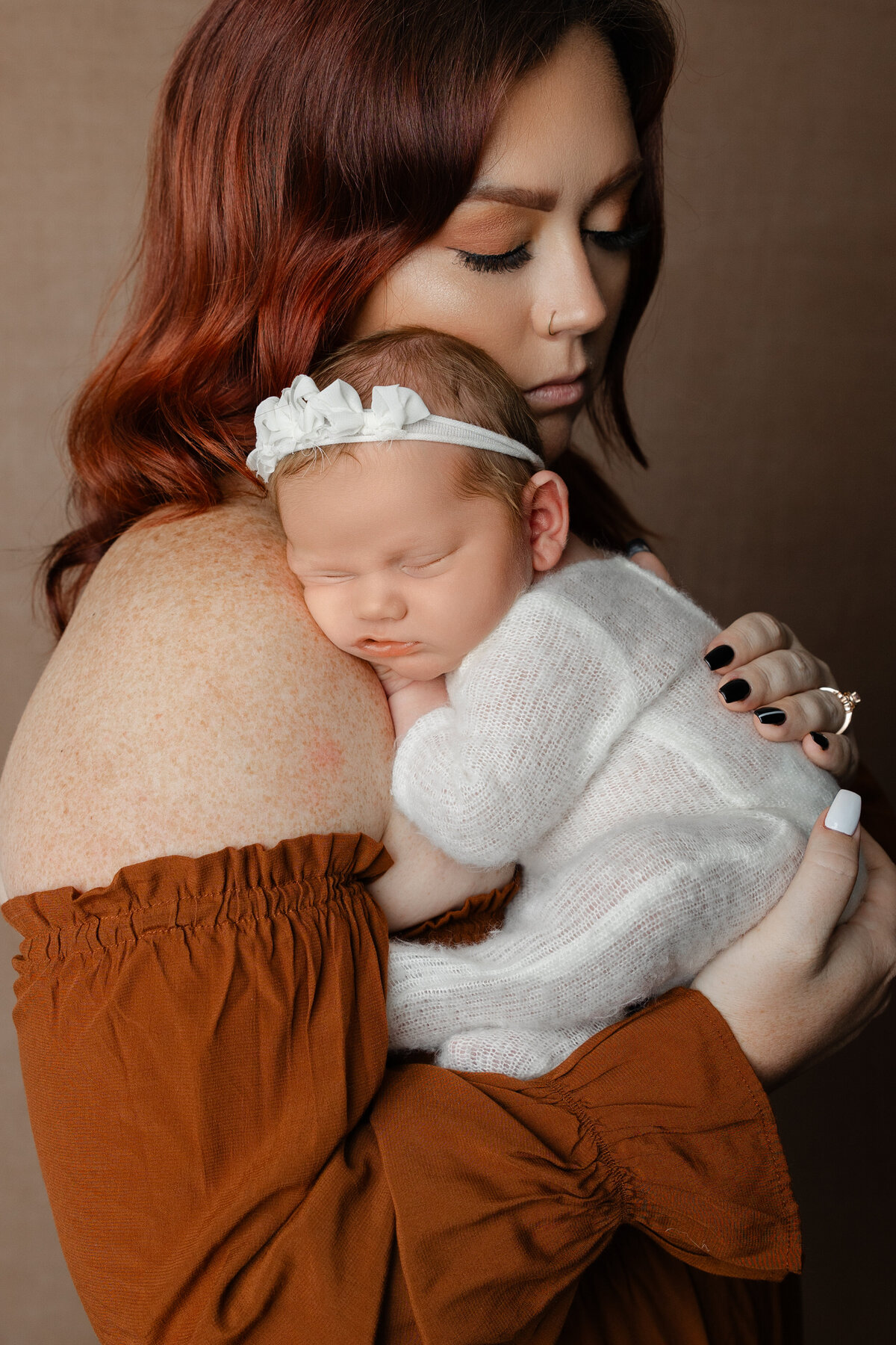 Mother embracing her sleeping newborn daughter dressed in a white knit outfit and floral headband during their studio session. Captured by Inland Empire newborn photographer Kaitlyn Dawn Photography.