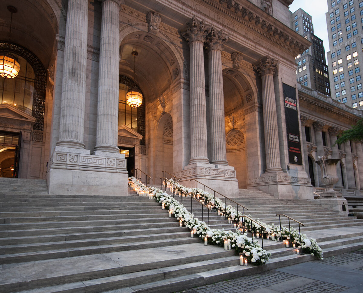 The image was taken at New York City, The New York Public Library by photographer Christian Oth. The grand entrance of the building is captured in the photo, showcasing tall columns and wide steps adorned with white floral arrangements and lit candles lining the steps. In the background, you can see evening lighting and adjacent city buildings. Additionally, East West Films were the videographers present at the location.