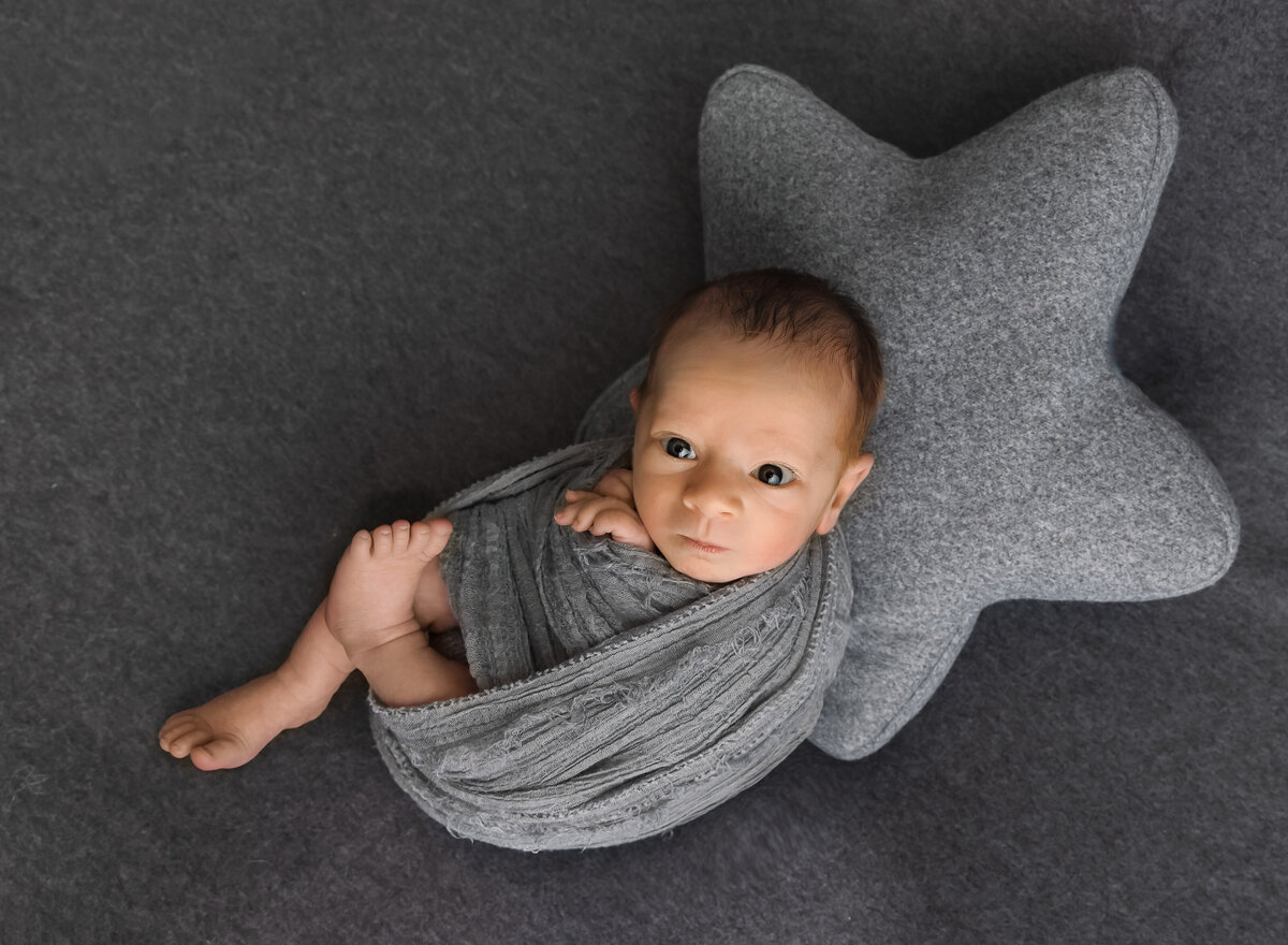 Baby boy is awake for his brooklyn ny newborn phtotoshoot. Baby is swaddled in grey with his legs and fingers peeking out of the swaddle. He is laying atop of a large felt star pillow.