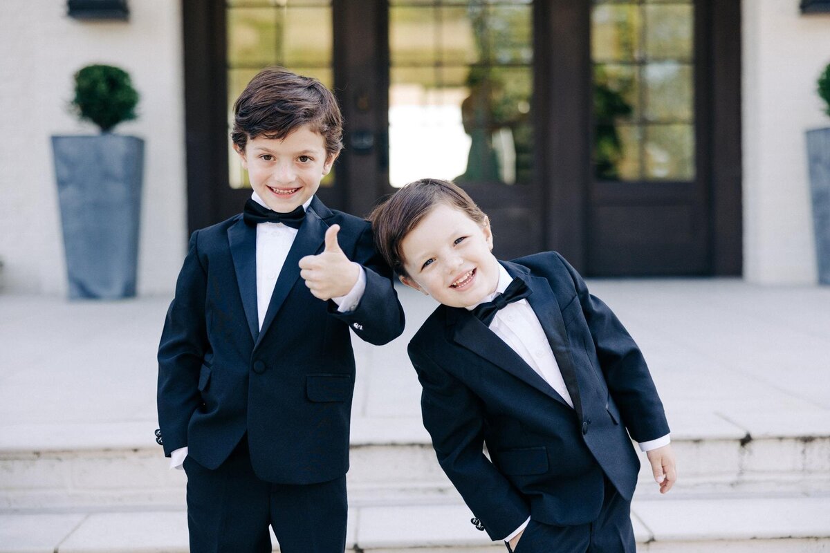 Two young boys in tuxedos smiling and gesturing, possibly at a formal event.