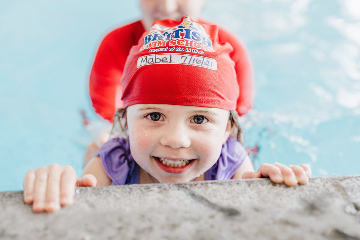 girl smiles at camera over the side of the pool while wearing. red bathing cap