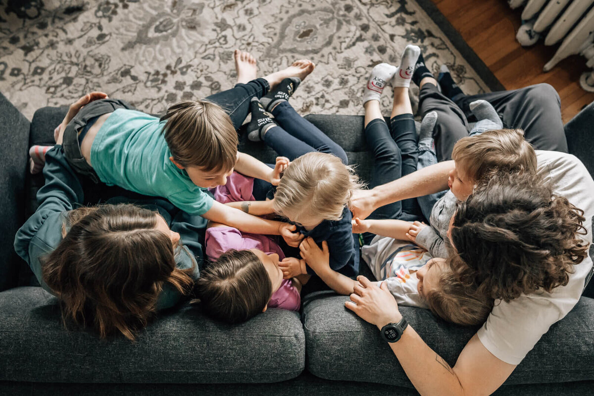 A photograph by Kate Simpson Photography, or a large family with small children cuddling on their couch, shot from above.