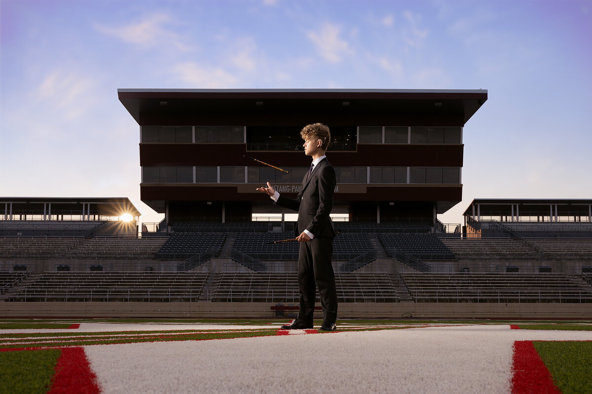 A senior in a suit confidently juggles a baton on the football field, captured by a Dallas senior photographer at Mustang-Panther Stadium.