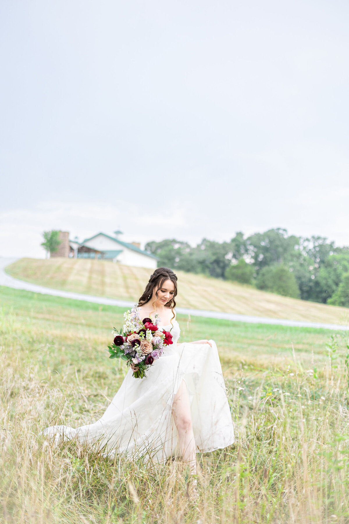 A bride holding a vibrant floral bouquet, walking gracefully through a serene countryside field with the Willow and Stone Events venue in the background in North Carolina