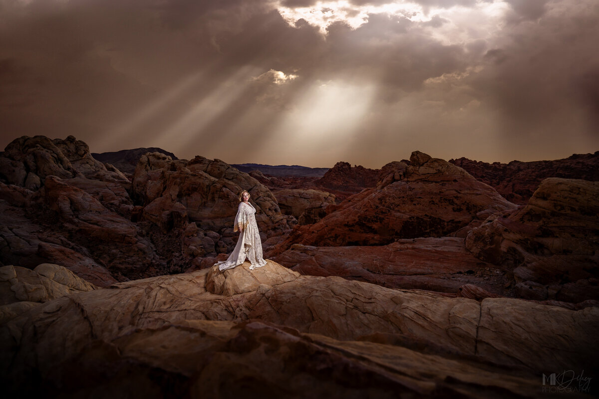 bold and glamorous over 50 beautiful woman photographed in valley of fire nevada, 50th birthday photography, elegant art deco dress, outdoor photography around las vegas, sunbeams shining on woman  barefoot on the rocks near cliffs