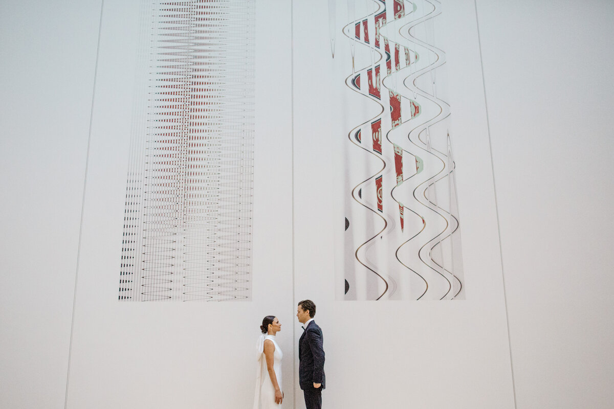 Wide angle view of bride and groom facing each other with large art feature as backdrop