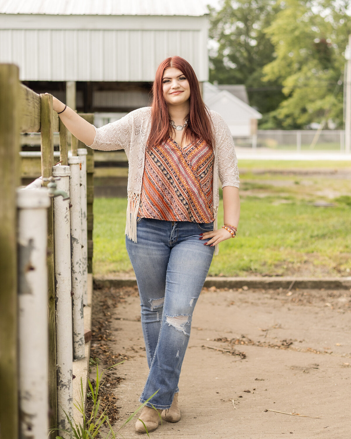 High school senior female with long red hair, wearing blue jeans, light tan smock over a patterned shirt, standing next to a fence with a building in the background smiling at the camera
