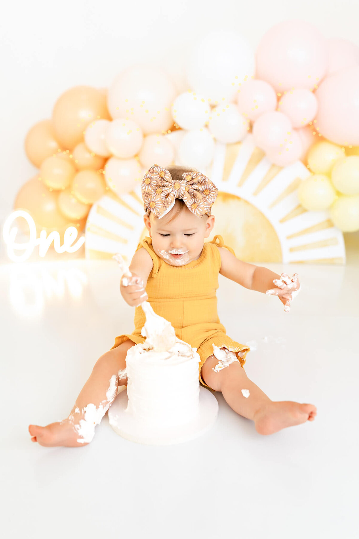 A toddler in a yellow outfit sits on the floor, playfully smashing a white cake. The background is decorated with pastel balloons and a sign that reads "One," indicating a first birthday celebration.